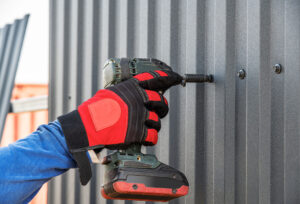PHOTO BY BELIEVE_IN_ME/© GETTY IMAGES A worker wearing glove drills a screw into a metal wall.