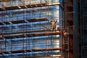 Photo by Tricky_Shark/(c) Shutterstock/Courtesy AT-PAC Worker installing scaffolding on building, showcasing the teamwork and safety equipment required for scaffold setup on construction site, focusing on safety harnesses and work platforms.