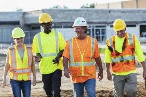 Photo (c) Getty Images A multiracial group of four construction industry workers is walking side by side through a construction site, conversing. The group includes a young Hispanic woman, a mid-adult African-American man, and two mature men.