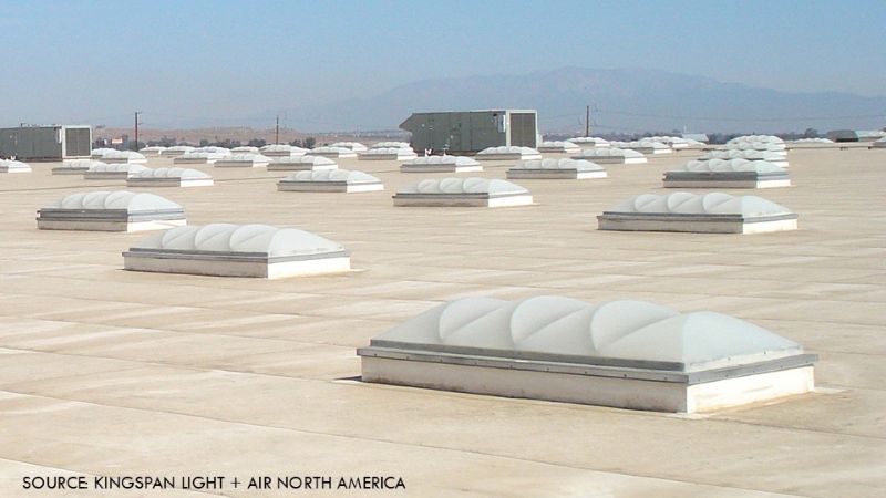 Photo shows a generic flat commercial roof covered with multiple evenly spaced dome skylights, with rooftop equipment and distant mountains visible in the background.