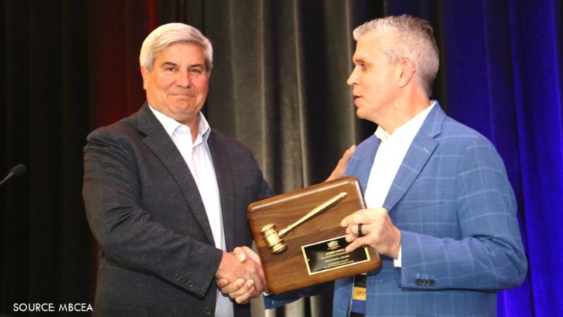 Two men shake hands as one presents a ceremonial gavel plaque during an MBCEA leadership transition.