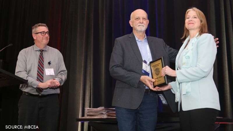 Award recipient accepts a plaque during a recognition ceremony as a presenter and emcee look on.