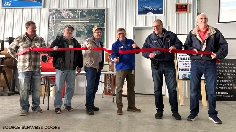Group of men holding a ribbon during a ribbon-cutting ceremony inside an aircraft hangar.