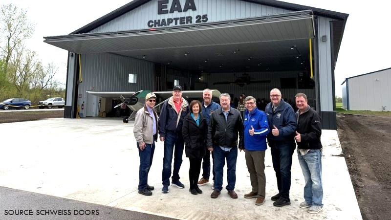 Group posing outside a metal aircraft hangar with an open bifold door and a small airplane visible inside.