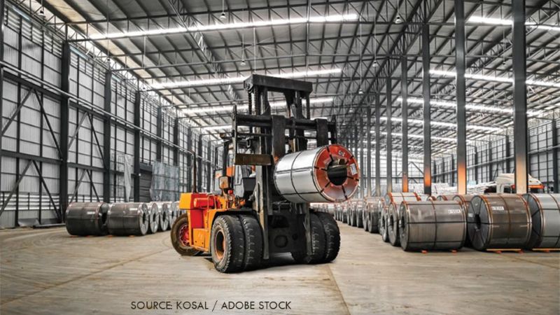Forklift transporting a large steel coil inside a warehouse with rows of stored metal coils.