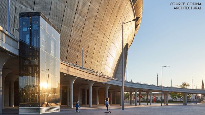 Exterior view of Puskas Arena showing its curved metal facade, glass elevator tower, and elevated pedestrian walkway at sunset.