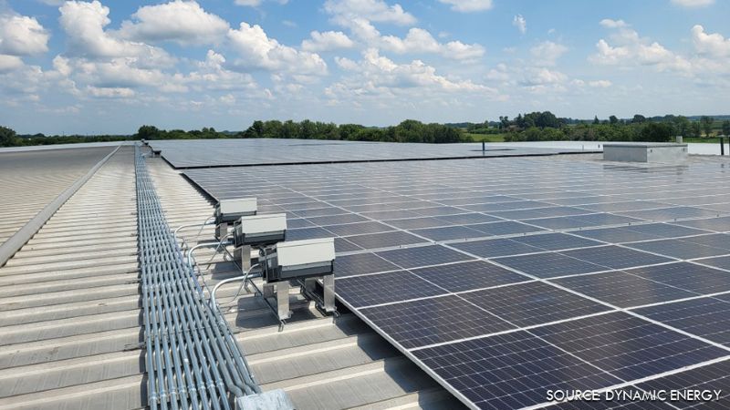 Rooftop solar array installed across a low-slope metal roof, with electrical equipment and conduit visible alongside the panels.