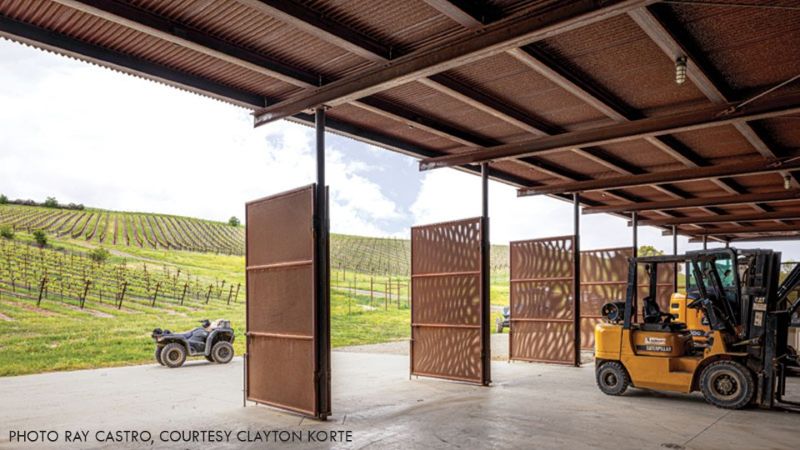 Interior of the Alma Rosa Vineyard barn showing open perforated metal doors, a forklift, and vineyard rows beyond.