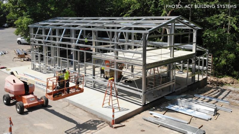 Two-story cold-formed steel building frame under construction, with workers on an aerial lift and building materials stacked nearby.
