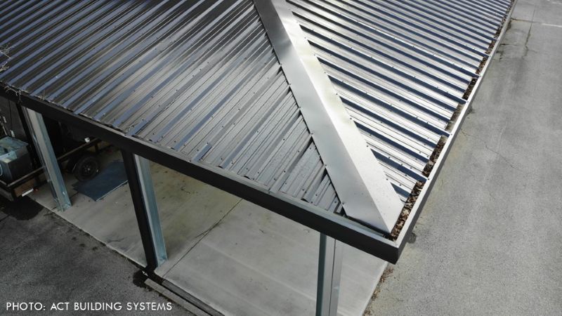 Overhead view of a sloped, ribbed metal roof with a central valley, supported by exposed steel columns over a concrete slab.