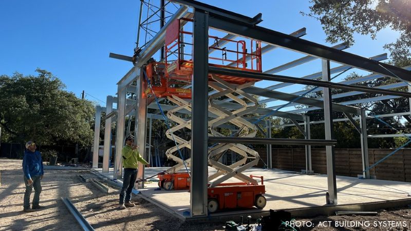 Cold-formed steel building frame under construction, with a scissor lift positioned inside the structure and two workers standing nearby.