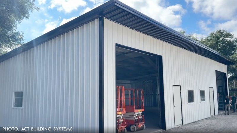 Completed steel building with white vertical ribbed metal wall panels, dark roof trim, windows, personnel doors, and large open overhead doorways.
