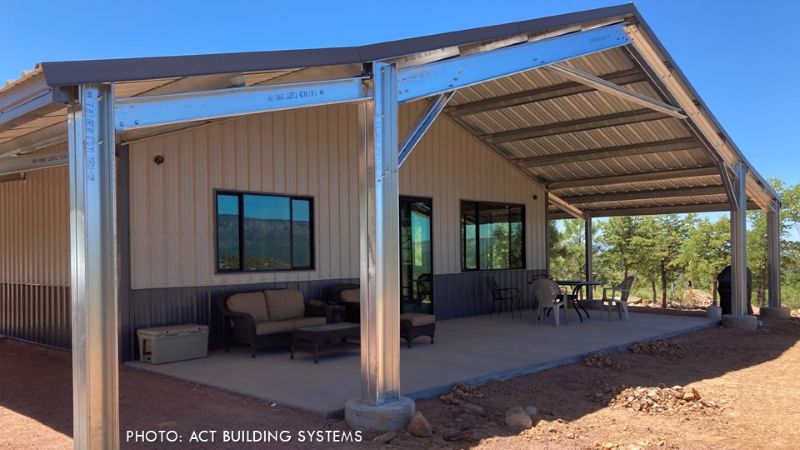 Covered porch of a steel-framed mountain home, featuring exposed steel columns and beams, metal roofing, outdoor seating, and wooded surroundings.